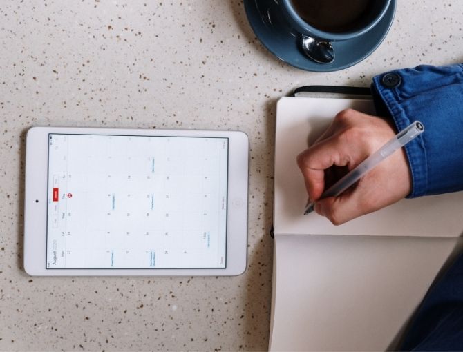 Person with pen over journal next to coffee mug and tablet with calendar
