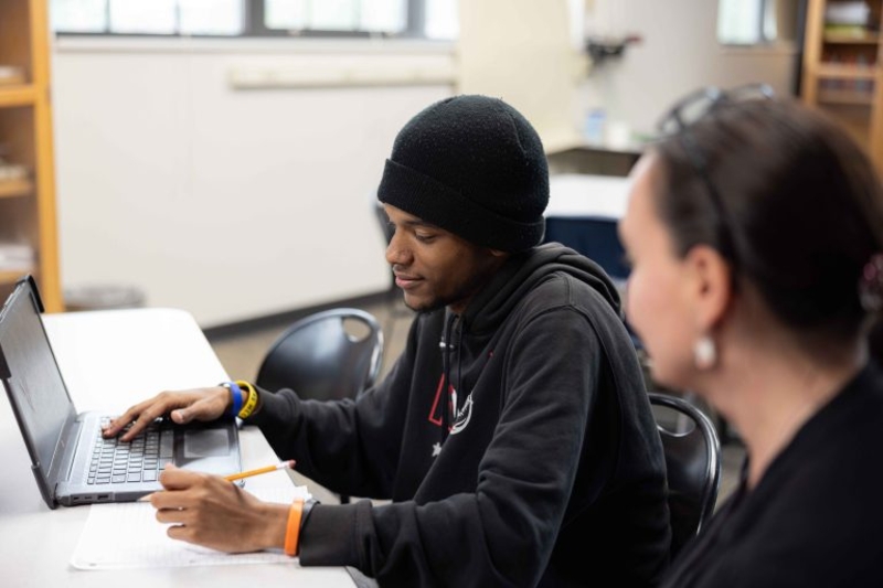 Teacher next to student holding pencil with one hand on laptop