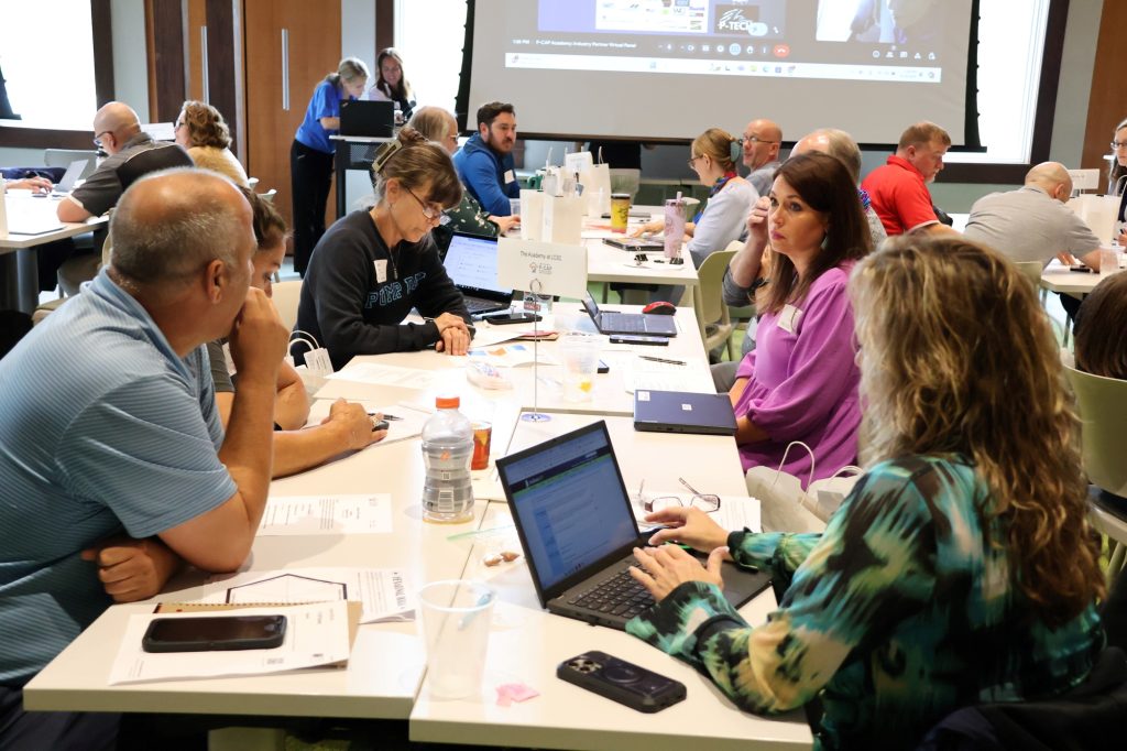 Participants sit in small groups at tables, working on laptops in a conference room
