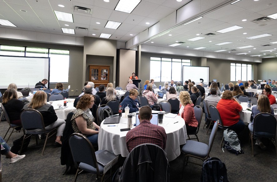 Large conference room with attendees seated at round tables, listening to a speaker at a podium near projection screens.