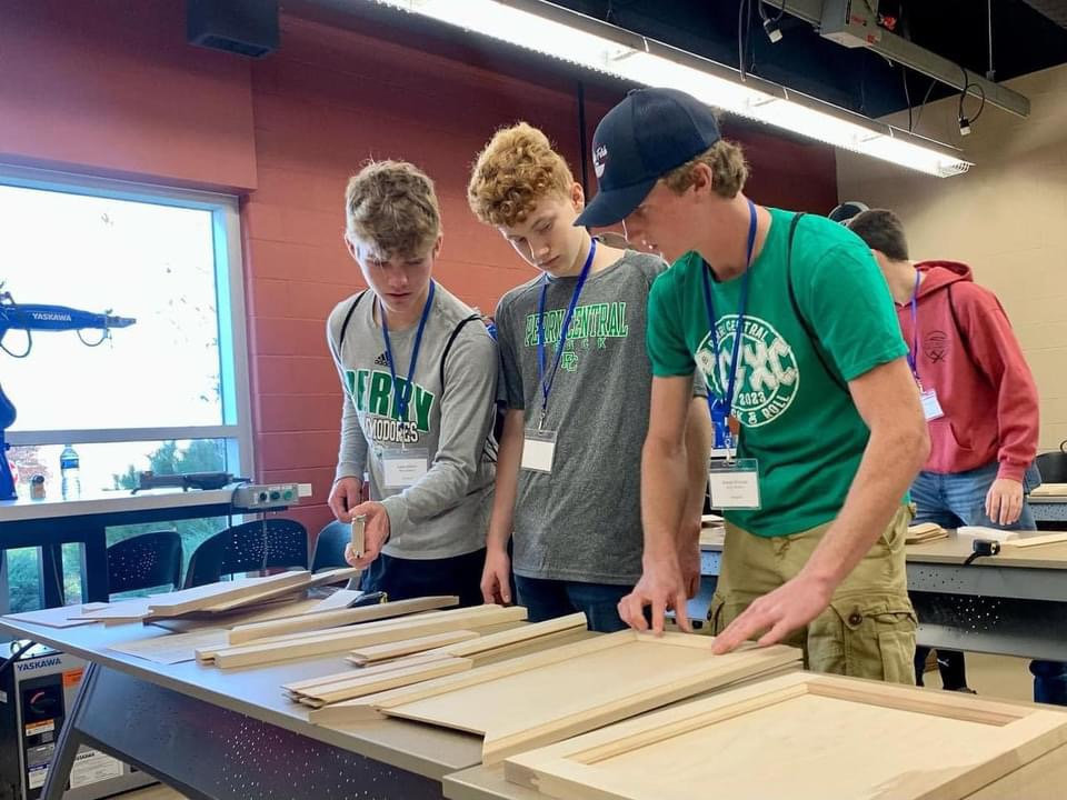 Three students wearing name badges examine wood pieces and panels laid out on tables in a workshop classroom.