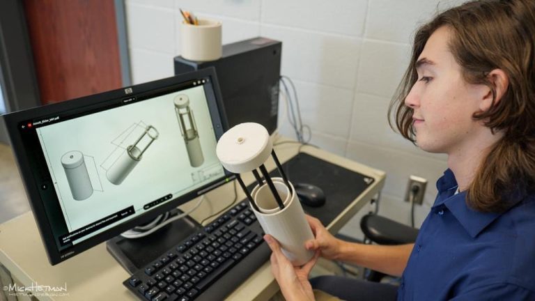 Person at a desk compares a cylindrical mechanical prototype in hand with a 3D CAD model displayed on a computer monitor.
