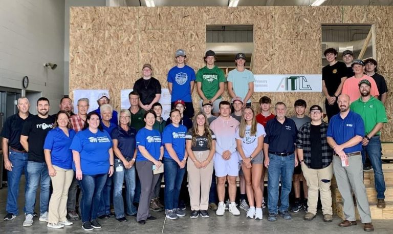 Large group of students and adults posing for a photo in front of a plywood-walled construction or workshop structure indoors.
