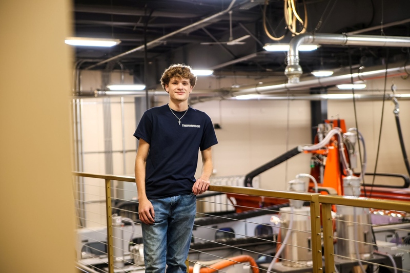 Young man standing by a railing inside an industrial workshop with exposed pipes and machinery in the background.