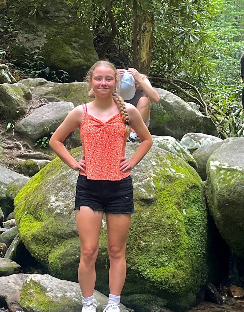 Person in an orange sleeveless top and black shorts stands with hands on hips on a moss-covered boulder in a wooded area.