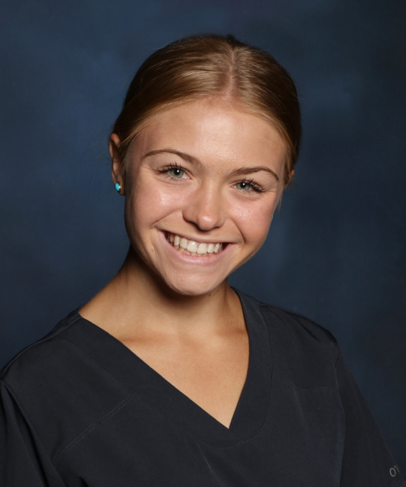 Studio headshot of a smiling woman with hair pulled back, wearing a dark V-neck top against a blue-gray background.