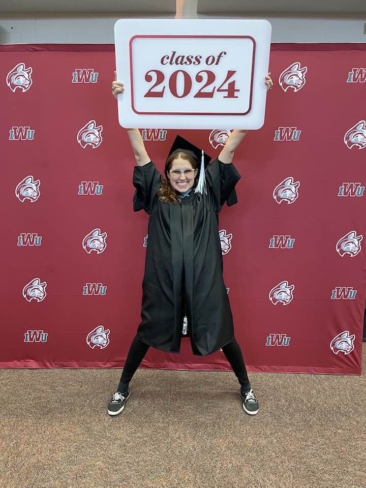 Graduate in cap and gown holds up a “class of 2024” sign in front of a red IWU step-and-repeat backdrop.