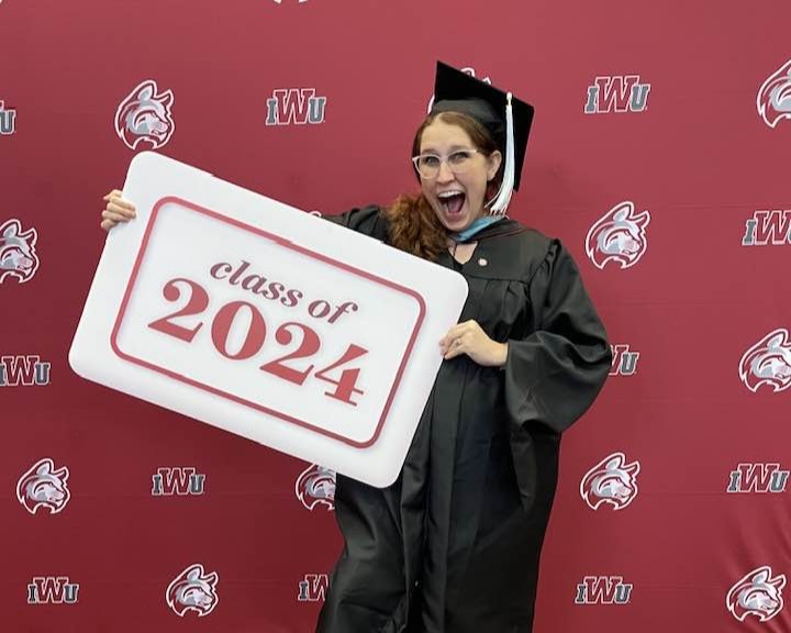 Smiling person in cap and gown holding "Class of 2024" sign with IWU backdrop