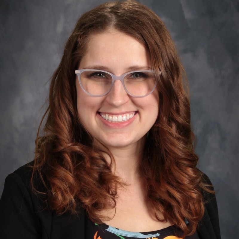 Smiling person with long wavy brown hair and light-colored glasses, photographed against a gray studio backdrop.