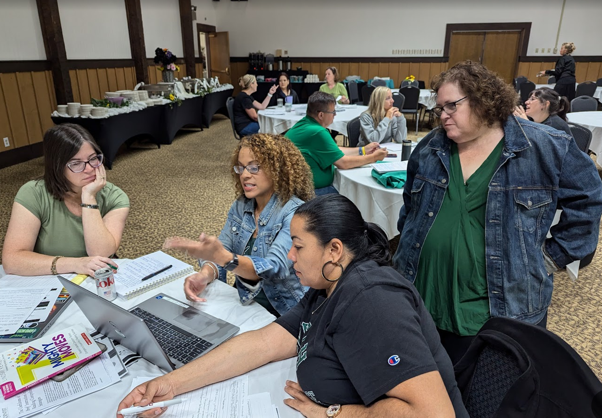 Three educators look at a workbook at a table with someone standing