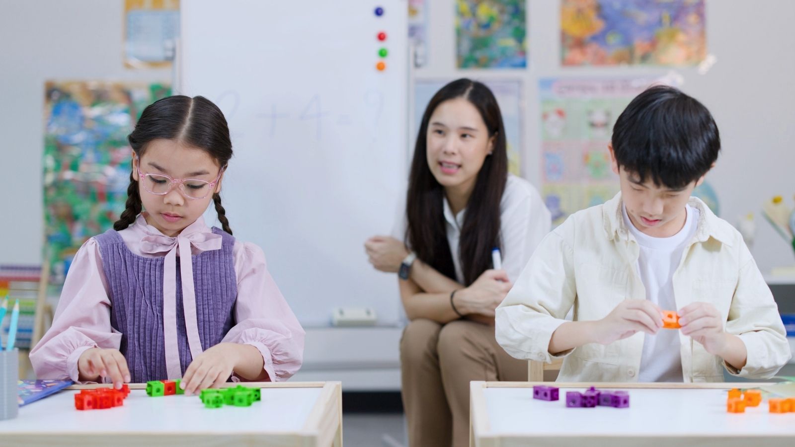 Two children build with colorful math cubes at their desks while a teacher watches in the background.