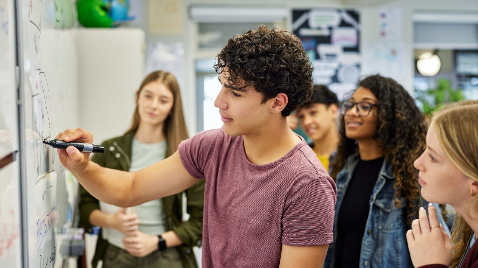 Student writes on a classroom whiteboard with a marker while classmates watch and discuss behind him.