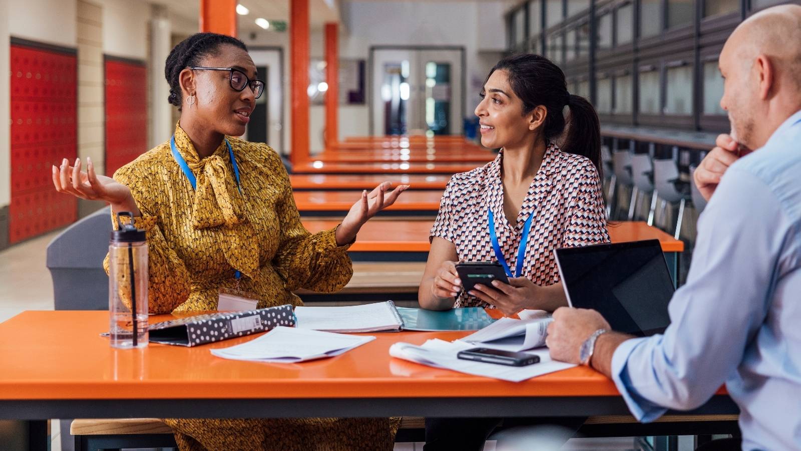 Three teachers sit at a table in a modern cafeteria, talking and reviewing notes with a laptop, calculator, and papers spread out.