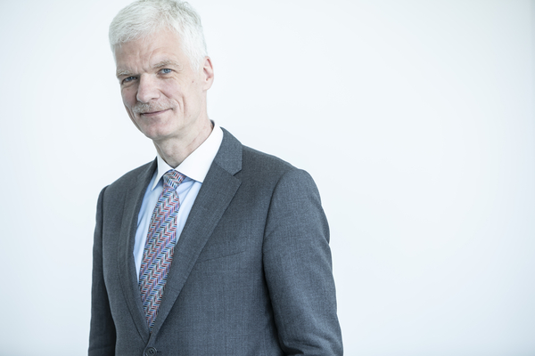 Man with short white hair wearing a gray suit and patterned tie, posing against a plain light background.